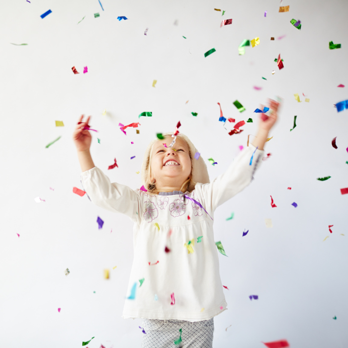 Joyful little girl, arms up, face skyward, throwing colourful confetti up in the air.